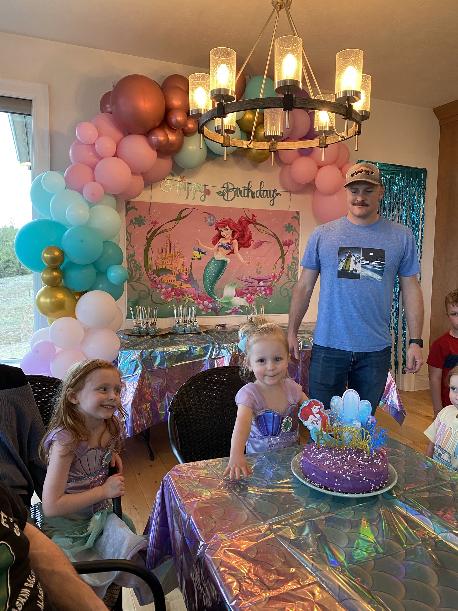 Guests celebrating as a pastel mermaid-themed birthday party with balloon garlands, dessert table, and colorful decorations.