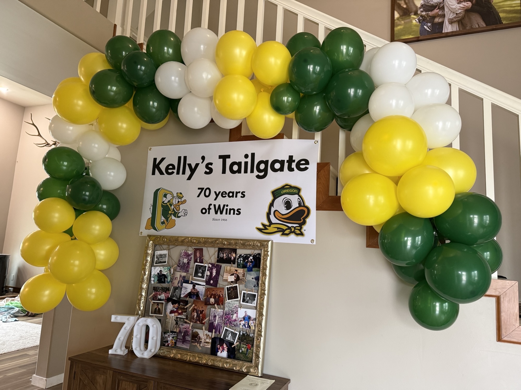Oregon Ducks-themed 70th birthday party backdrop with a green and yellow balloon garland, custom tailgate sign, and photo display.