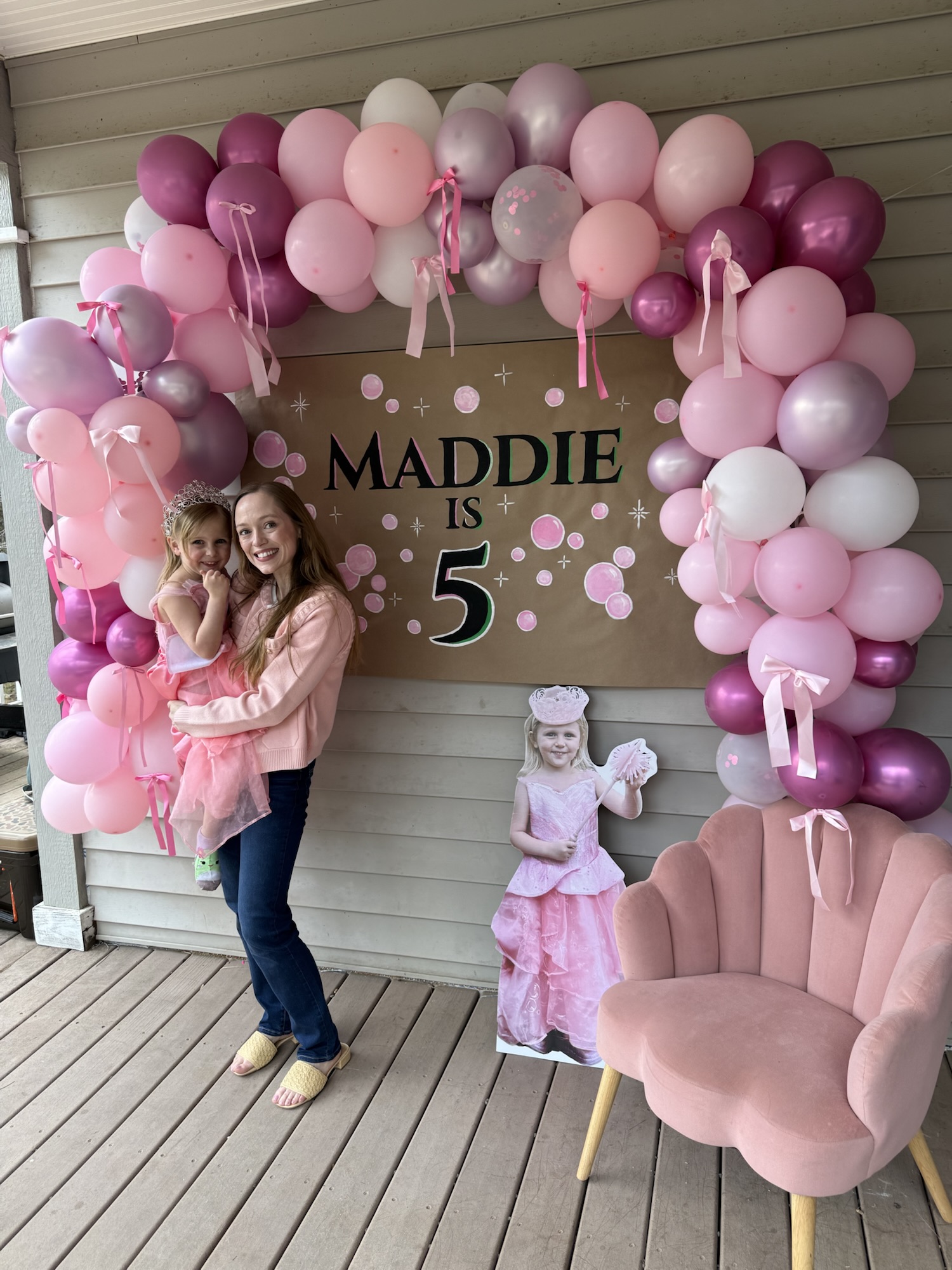 Mom hugging her daughter in front of a pink balloon arch and fifth birthday party backdrop.