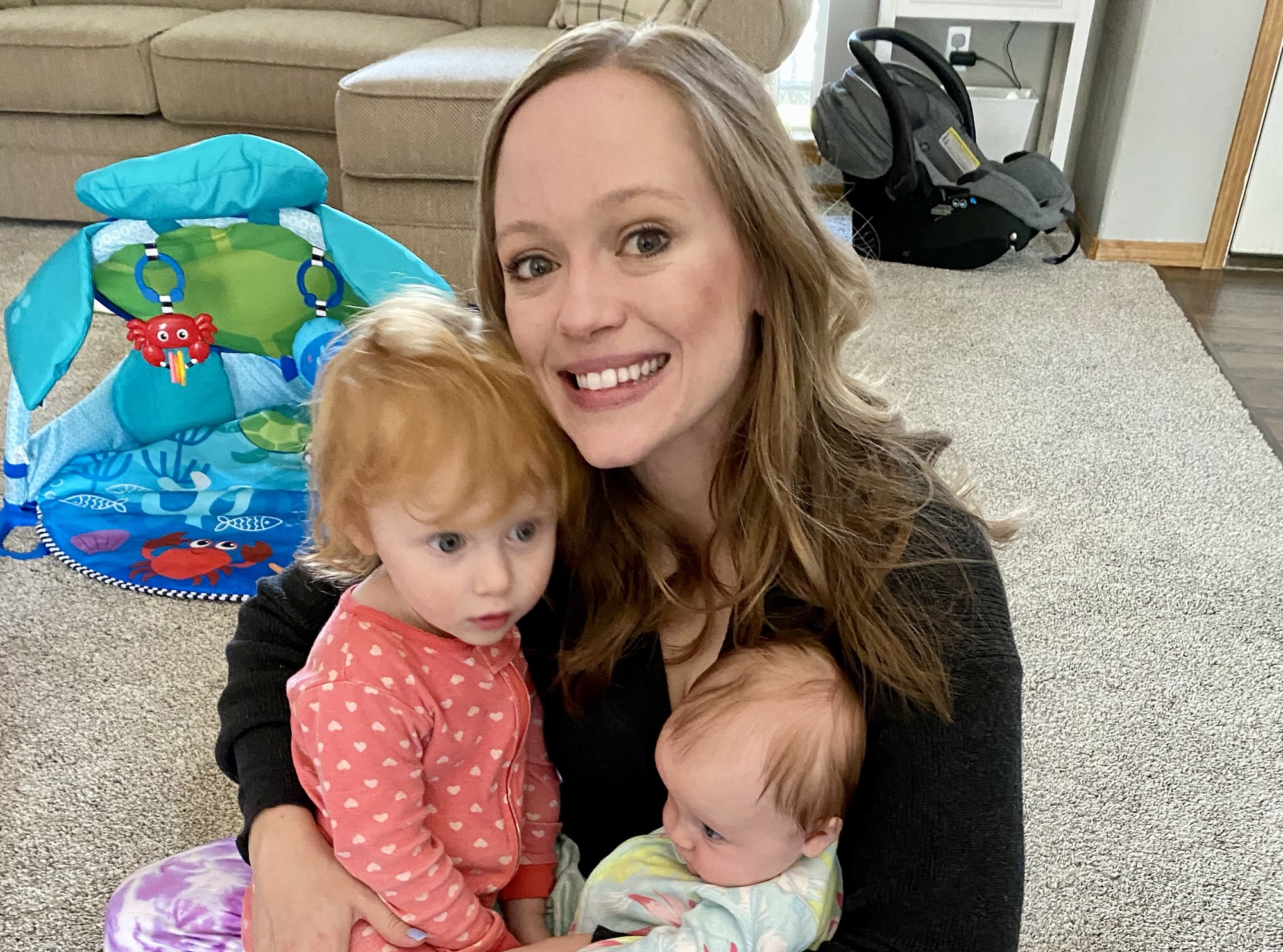 Mom smiling while holding her two young children in a living room with toys.