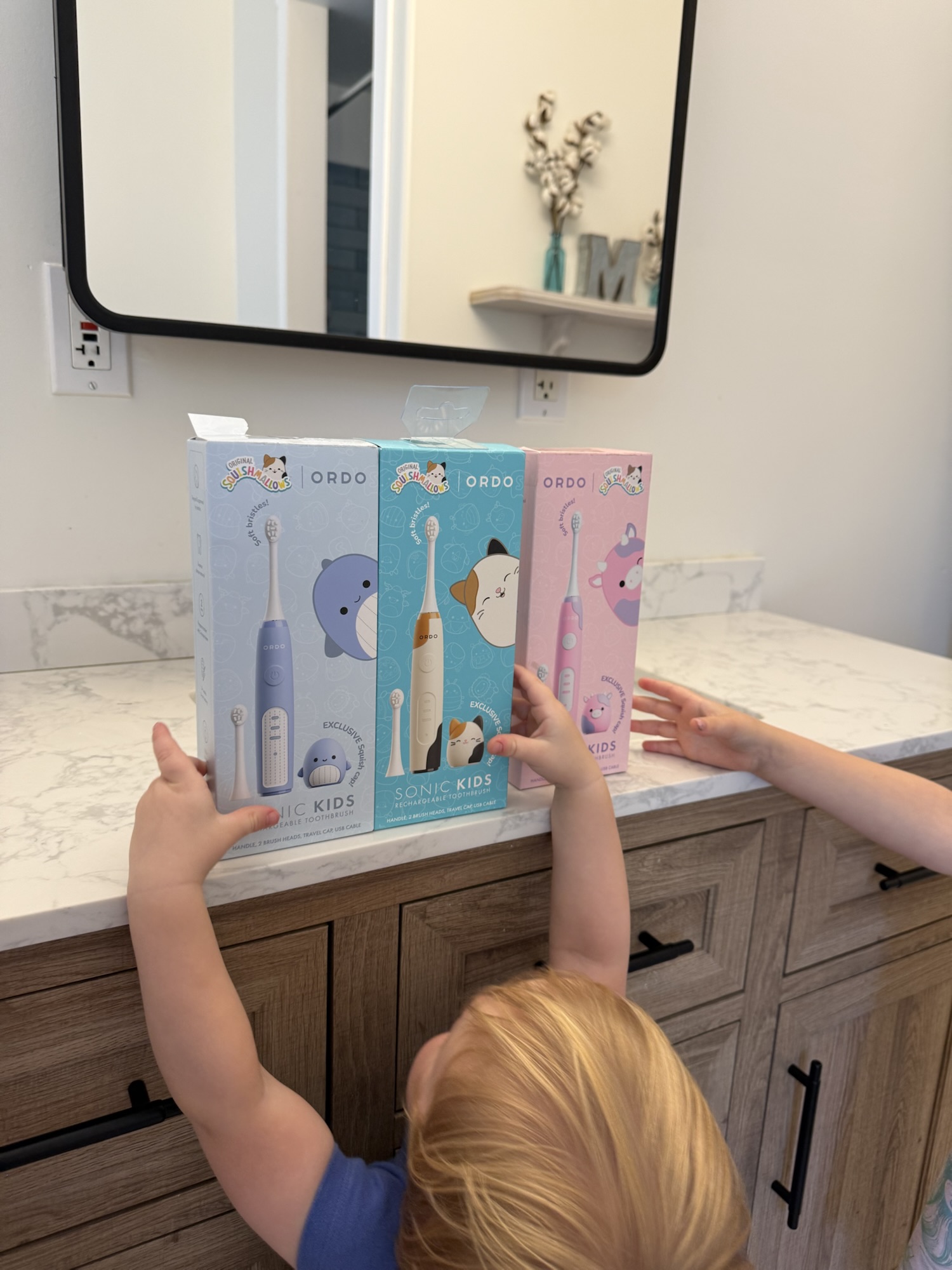Toddler reaching for Ordo Squishmallows electric toothbrush boxes before unboxing.